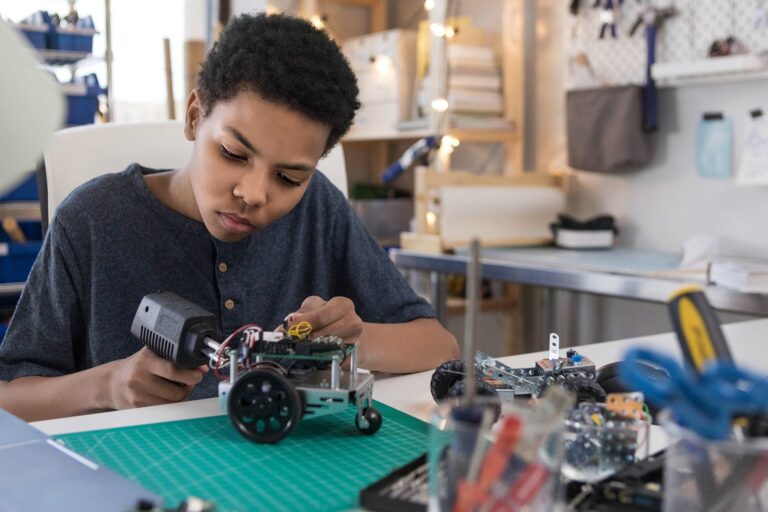 A serious teen boy uses a soldering gun to connect wires as he builds a robot at home.